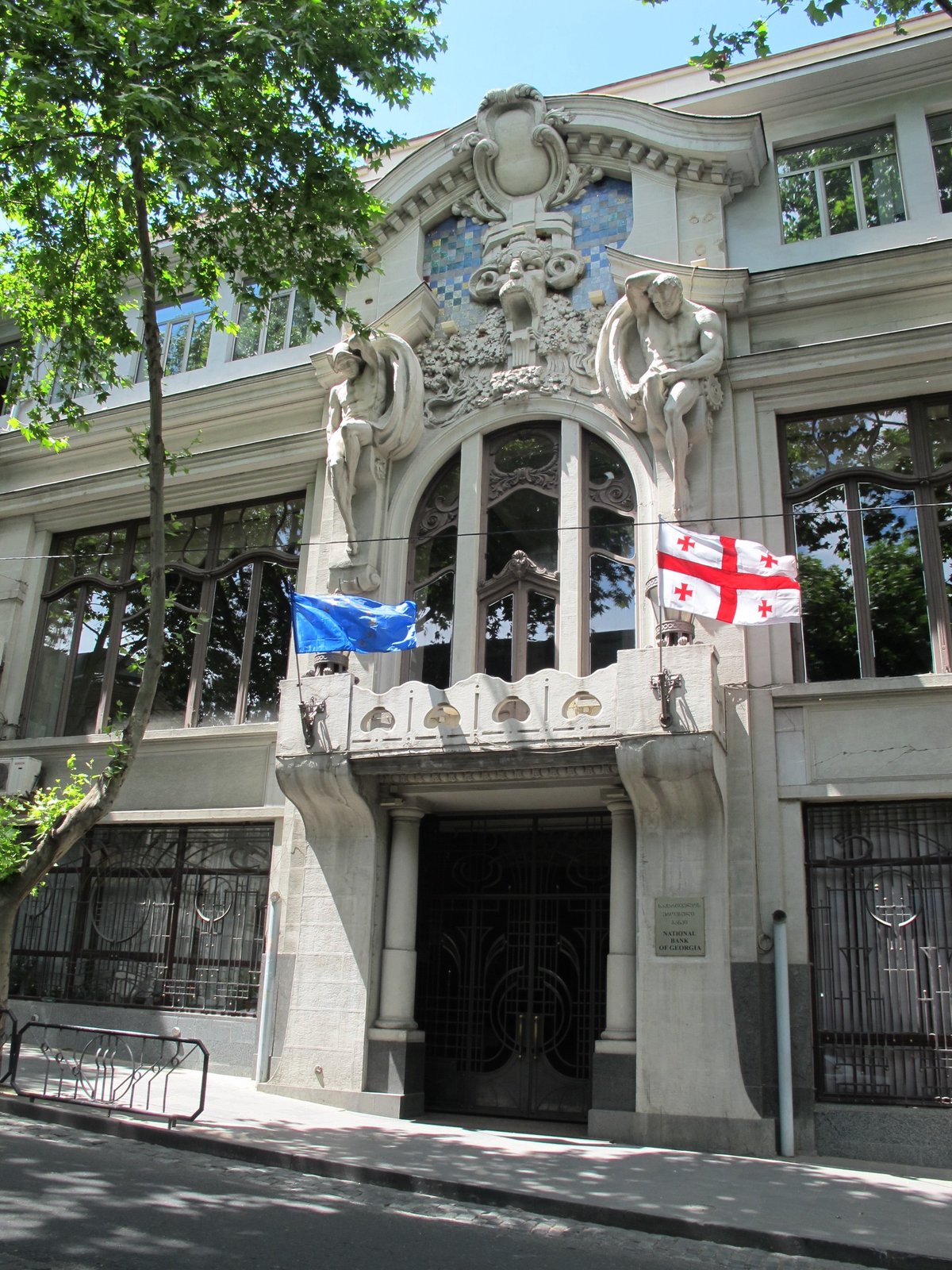 Art Nouveau facade of the former National Bank of Georgia headquarters at 3 Giorgi Leonidze Street, Tbilisi, with sculptural figures flanking a central window and the Georgian flag flying above the entrance