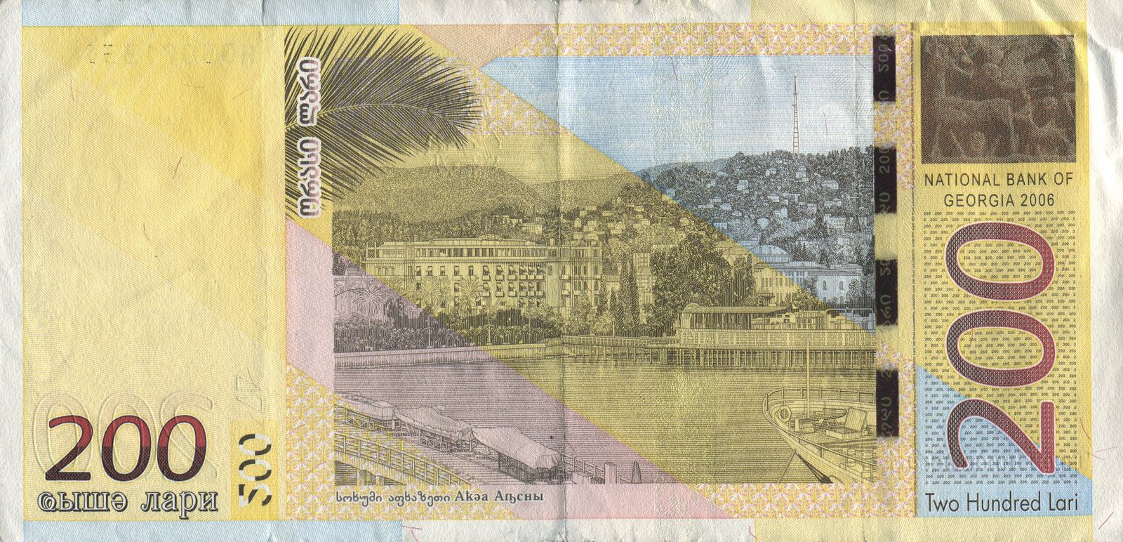 Reverse of the 200-lari Georgian banknote, showing the curving waterfront of Sukhumi with palm trees in the foreground, the long nineteenth-century embankment of white hotels, hills and a radio tower behind, a dark sandstone bas-relief fragment from Tsebelda inset at upper right, against a pale yellow ground, with denomination "200" markers at lower corners and trilingual Georgian-Abkhaz-English denomination text